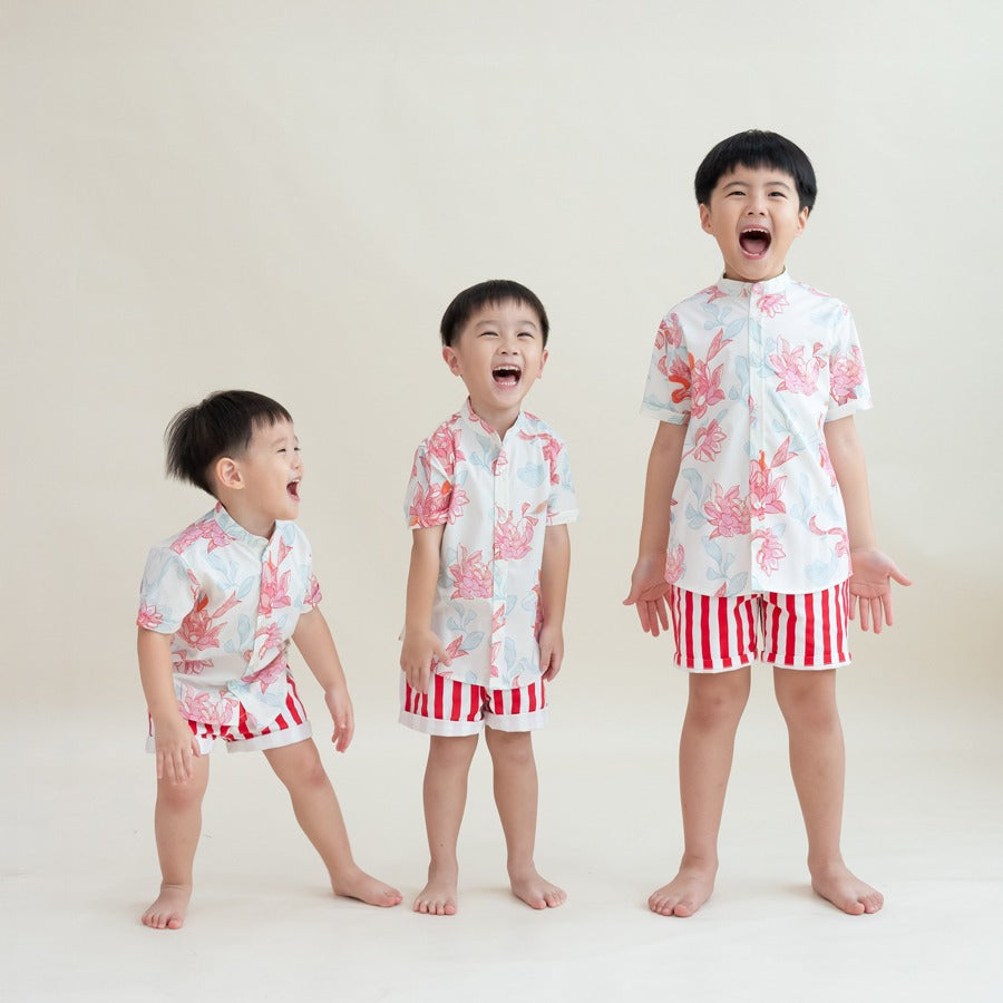 Three boys dressed in matching Lotus Collection outfits stand in a row, bursting into laughter. Their mandarin-collar shirts are covered in coral and pastel lotus illustrations, complemented by bold red-and-white striped shorts. The joyful energy of the m