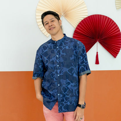 A male model wearing a navy blue chambray floral short-sleeve shirt from the Le Petit Society x Sheila Sim CNY 2026 collection stands casually with one hand in his pocket in front of a two-tone orange and white wall decorated with red and cream paper fans