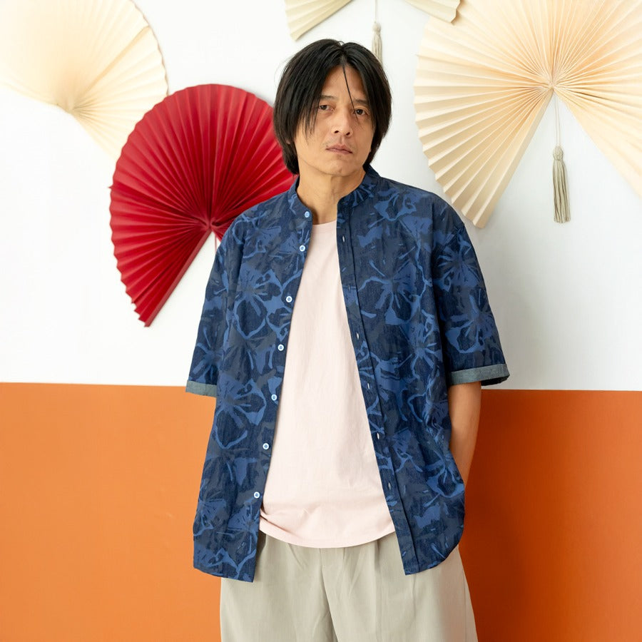 A male model wearing a navy blue chambray floral short-sleeve shirt from the Le Petit Society x Sheila Sim CNY 2026 collection stands with his hands in his pockets in front of a two-tone orange and white wall decorated with red and cream paper fans. He fa