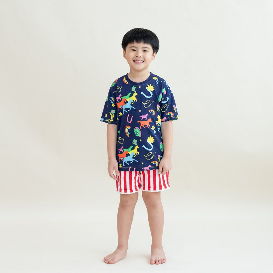 A young boy wearing a navy blue T-shirt with colourful horse and lucky symbol prints paired with red-and-white vertical striped shorts from the Le Petit Society x Sheila Sim CNY 2026 collection stands smiling against a soft cream background.