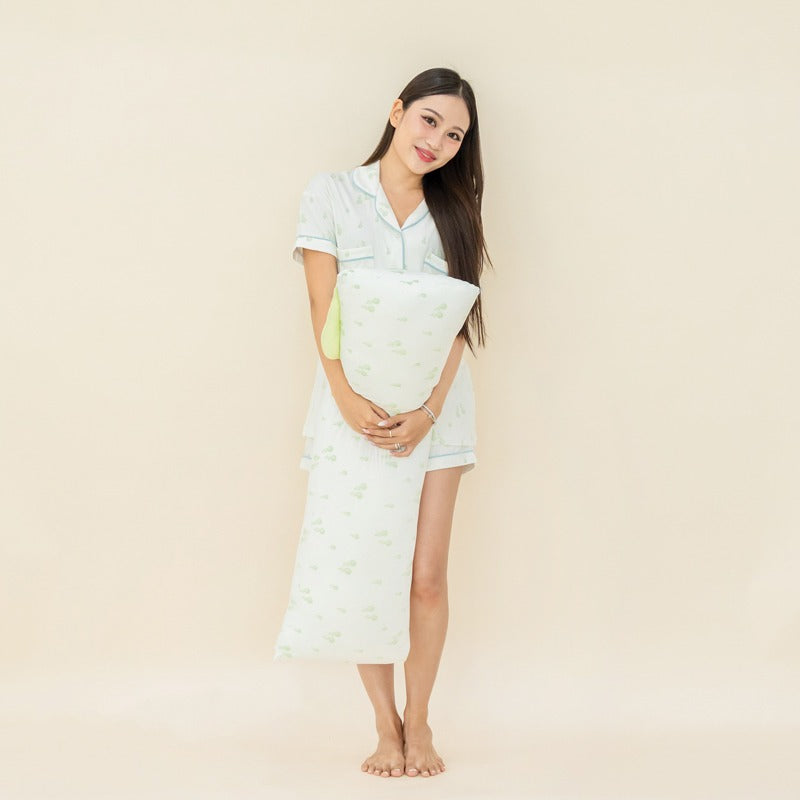An adult woman with a matching pyjamas set and pillow from Le Petit Society, with both featuring a print of green pears.