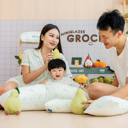 A family of three — mother, father, and baby boy — all wearing matching sleepwear and with pillows featuring a green pear print from Le Petit Society.