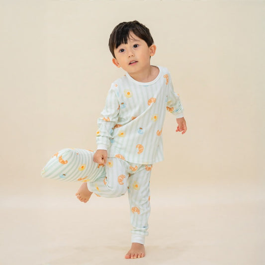 A little boy wearing a pyjama set and holding a matching pillow from Le Petit Society. These pieces feature pastel green stripes and a print of breakfast foo.