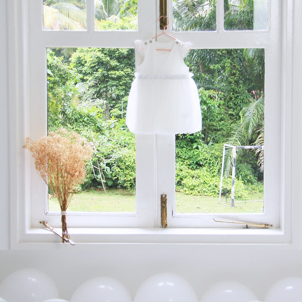 A white baby bubble dress with a tulle skirt and a pair of flutter sleeves is hung on a white window sill of a wedding venue overlooking lush greenery.