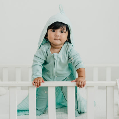A baby wearing a mint green organic zip sleepsuit in leaf print from Le Petit Society.