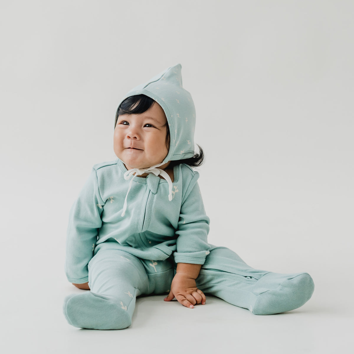 A baby wearing a matching organic mint blue bonnet hat and zip sleepsuit in leaf print from Le Petit Society.