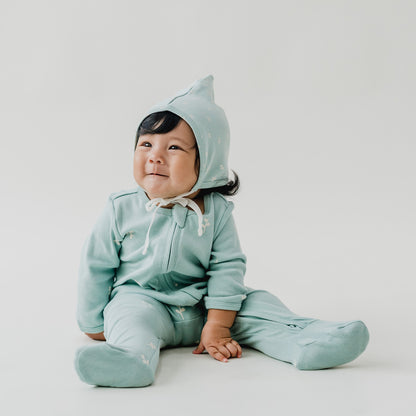 A baby wearing a matching organic mint blue bonnet hat and zip sleepsuit in leaf print from Le Petit Society.