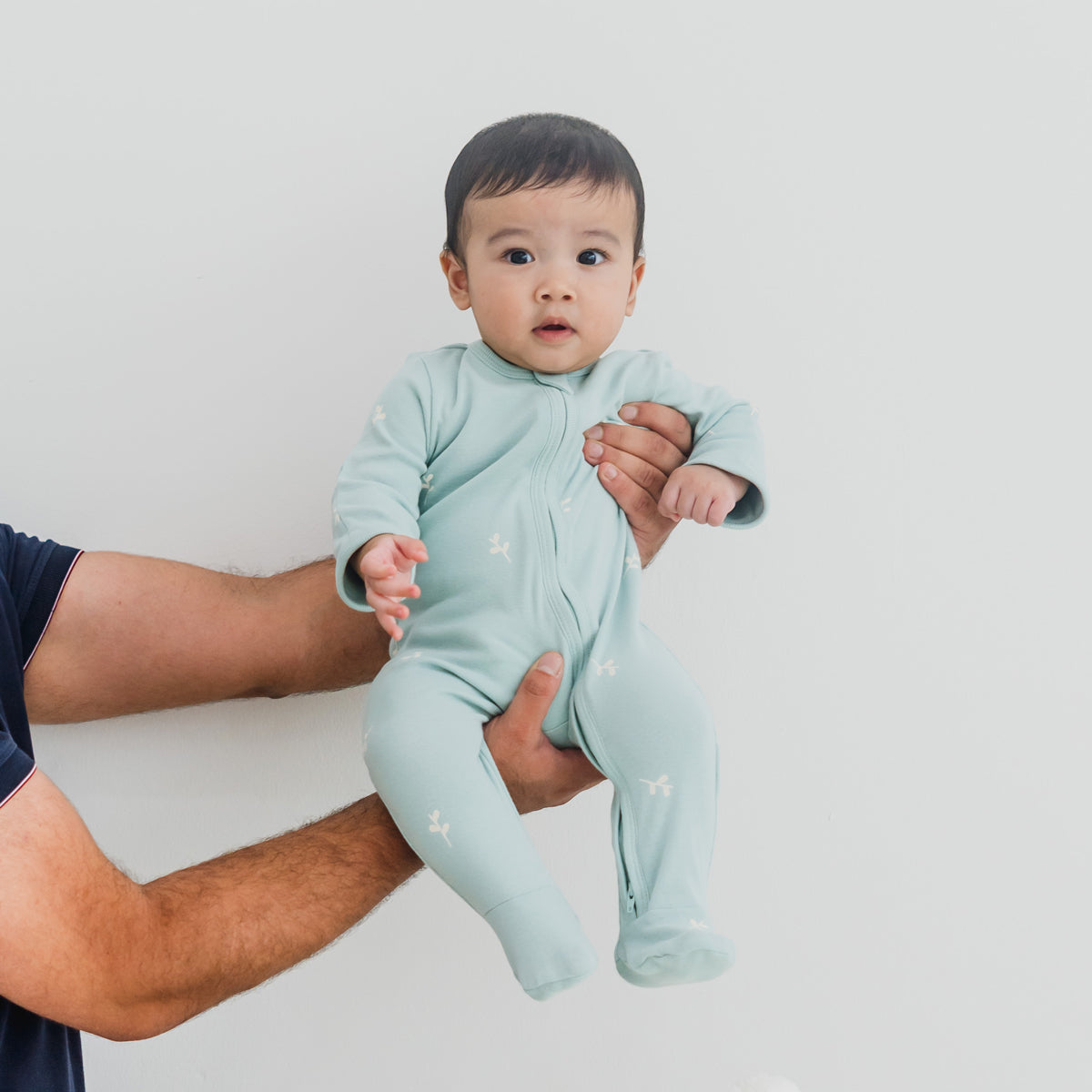 A baby wearing a mint blue organic zip sleepsuit in leaf print from Le Petit Society.