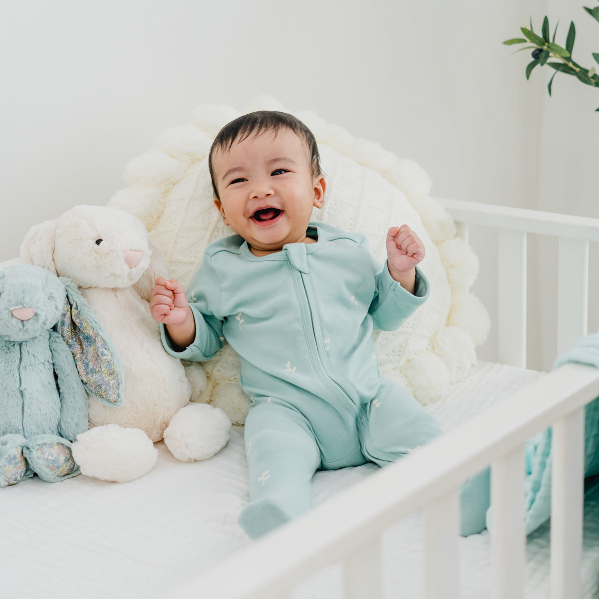 A happy baby wearing a mint blue organic zip sleepsuit in leaf print from Le Petit Society.