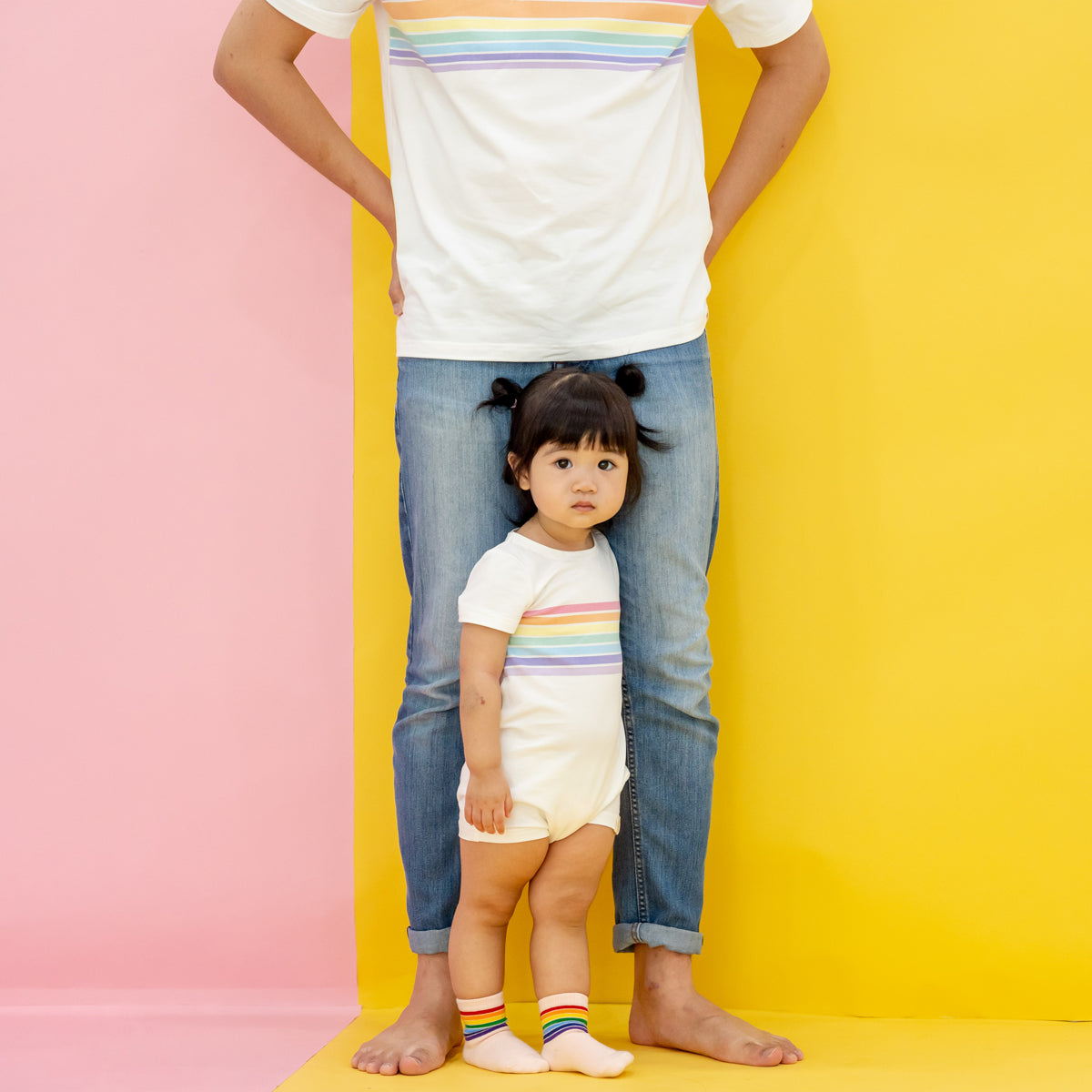 A toddler and father in matching outfits consisting of a white romper and white tee in the same pastel rainbow print from Le Petit Society's rainbow series. 