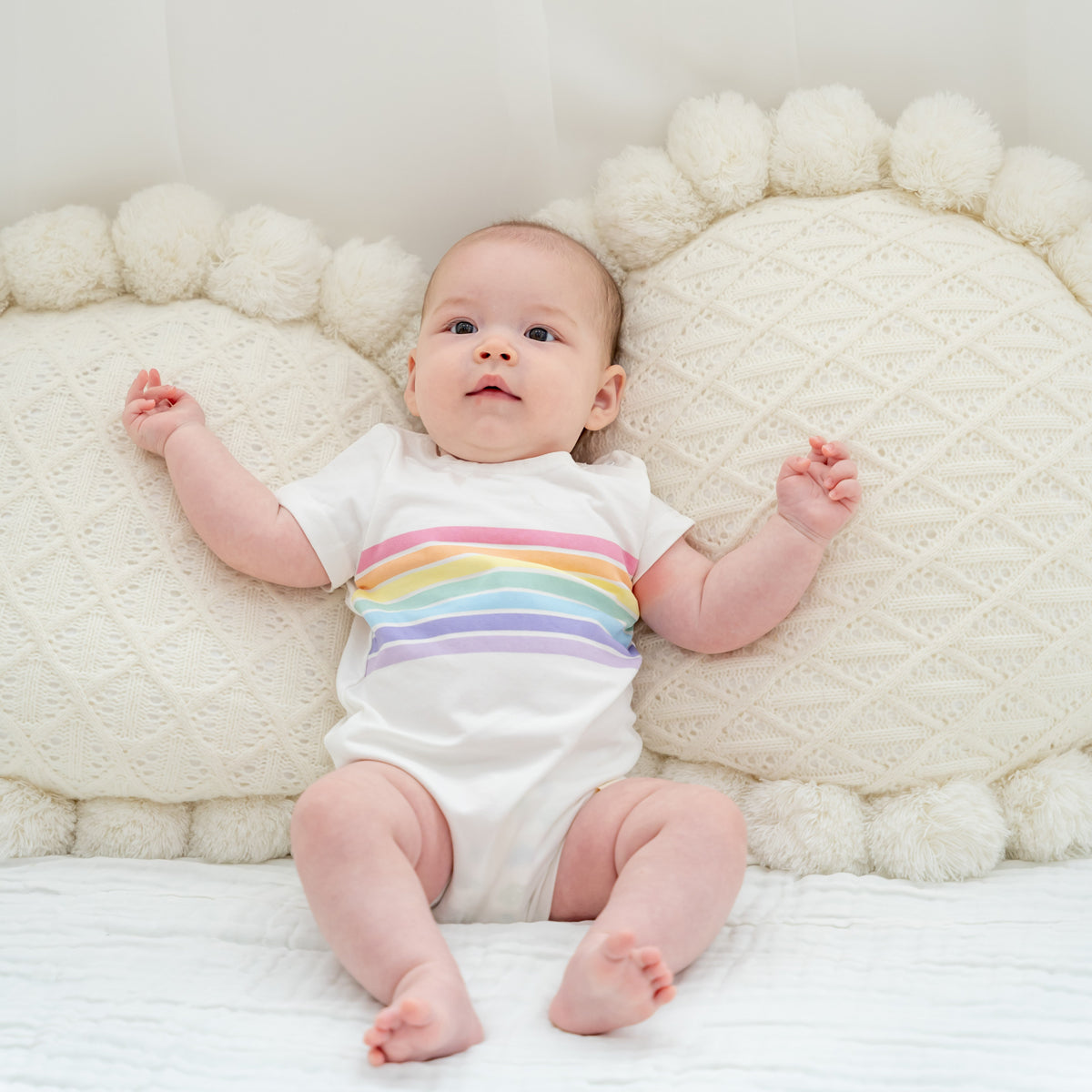 A baby wearing a white romper with a pastel rainbow print from Le Petit Society's rainbow series.