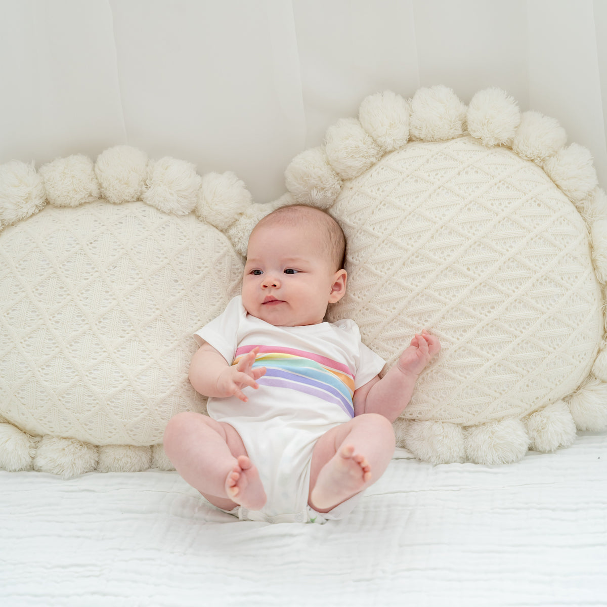 A baby wearing a white romper with a pastel rainbow print from Le Petit Society's rainbow series.