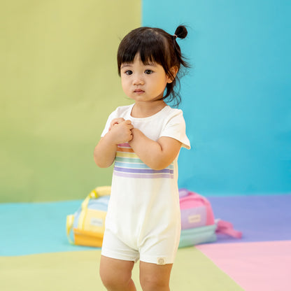 A toddler girl wearing a white romper with a pastel rainbow print from Le Petit Society's rainbow series.