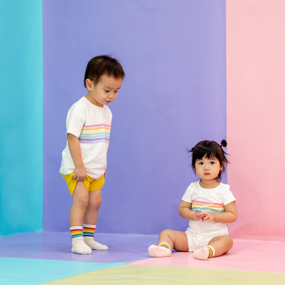 Two toddlers wearing matching outfits consisting of a white tee and white romper in the same pastel rainbow print from Le Petit Society's rainbow series.