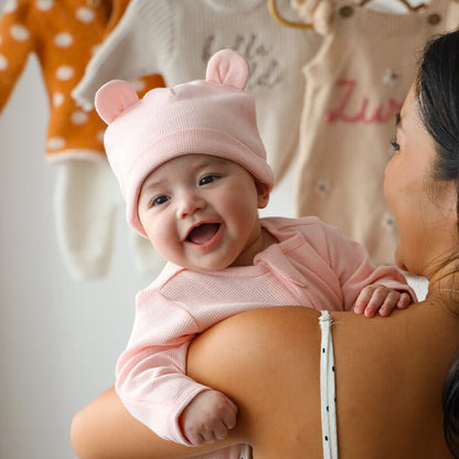 A baby wearing an organic bear hat and zip sleepsuit in pink by Le Petit Society.
