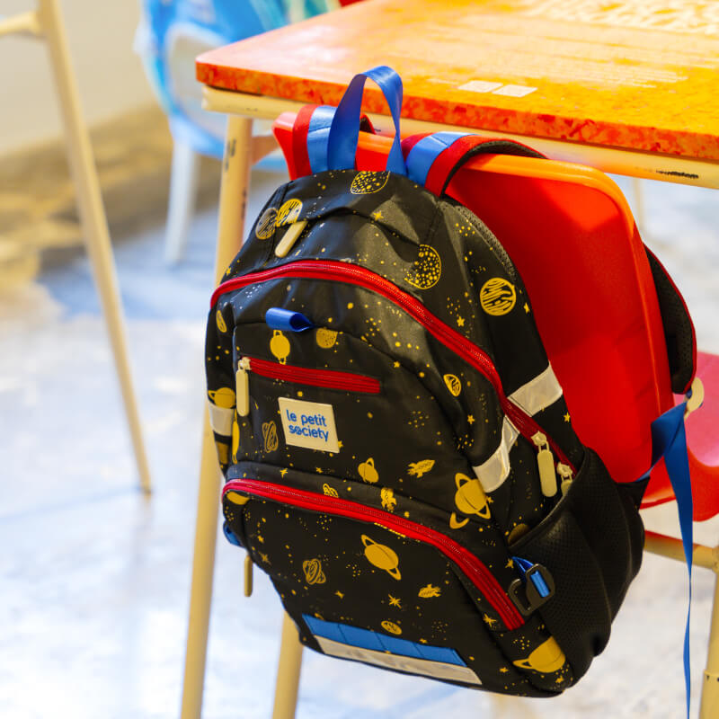 The LPS kids single pocket school bag hanging on to a classroom chair with its red and blue shoulder straps.