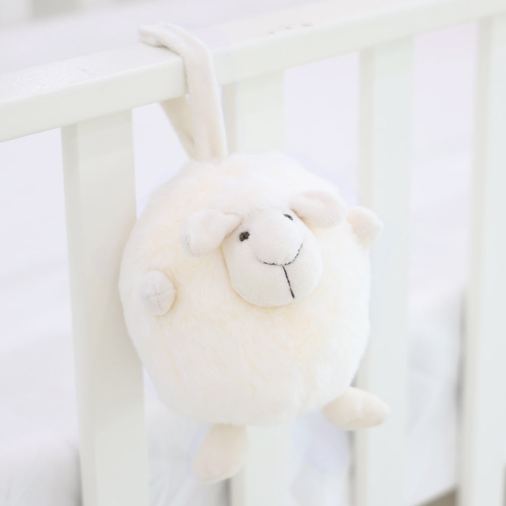 A round and plush sheep toy in cream white by Le Petit Society hangs off the frame of a baby cot.