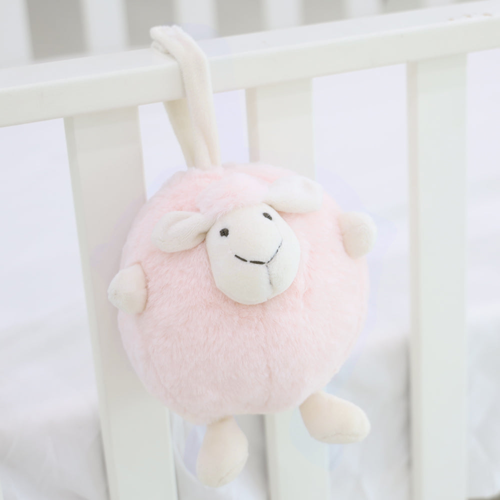 A round and plush sheep toy in soft pink by Le Petit Society hangs off the frame of a baby cot.