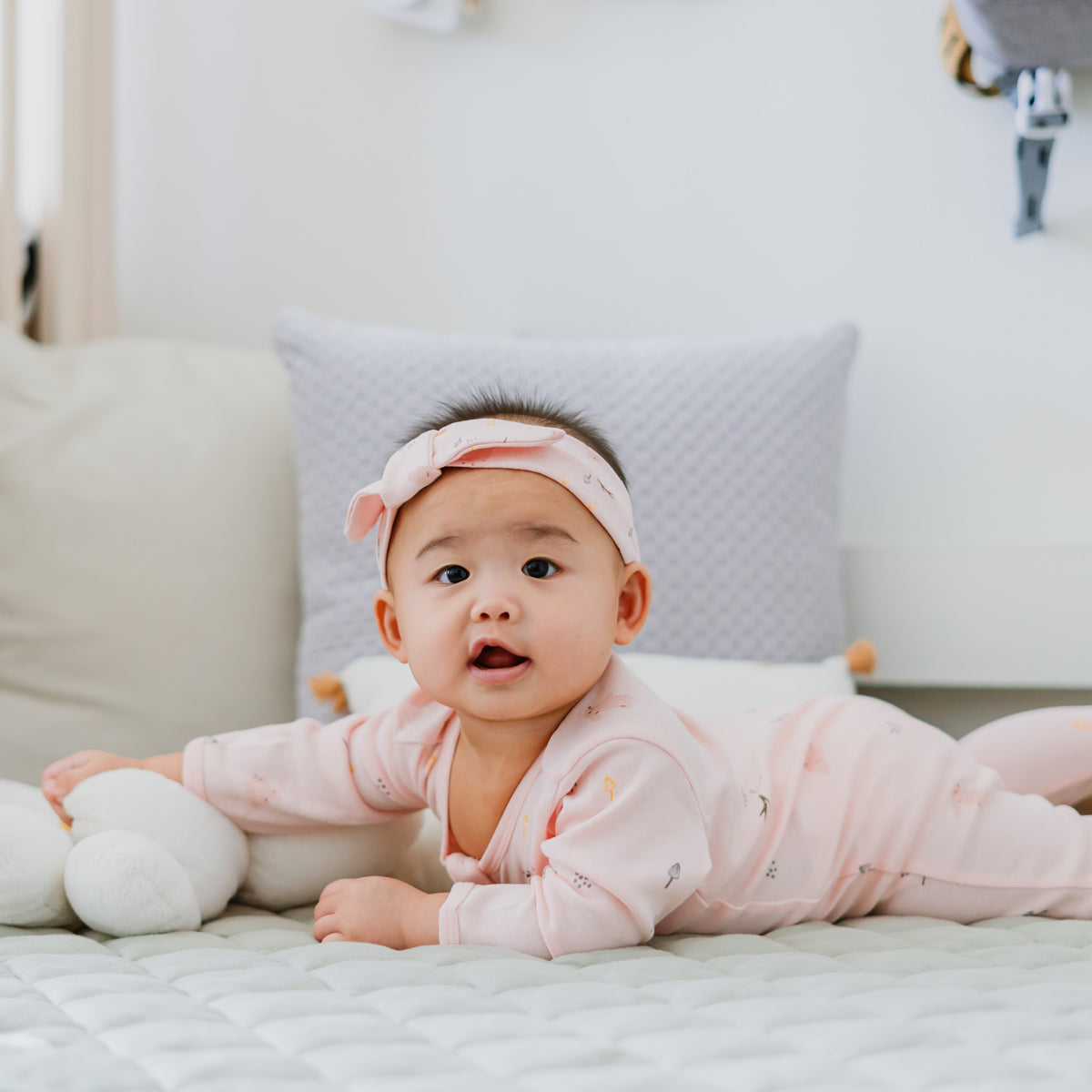 A baby wearing a pink zip sleepsuit and bow headband in a rabbit print by Le Petit Society,