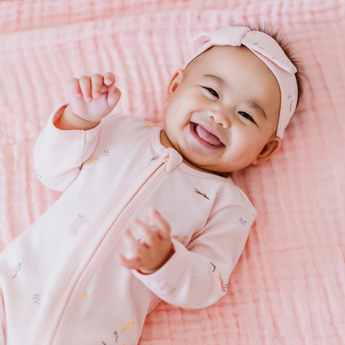 A baby wearing a pink zip sleepsuit and bow headband in a rabbit print by Le Petit Society,