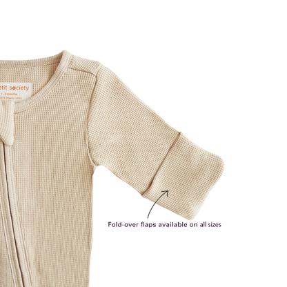 A baby organic zip sleepsuit in beige by Le Petit Society, with fold over flaps at the sleeves to cover a baby's hands, acting as a substitute for mittens.