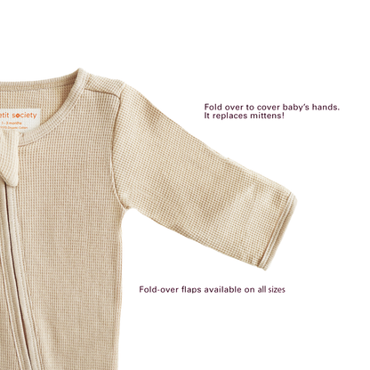A baby organic zip sleepsuit in beige by Le Petit Society, with fold over flaps at the sleeves to cover a baby's hands, acting as a substitute for mittens.

