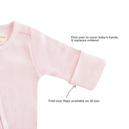 A baby organic zip sleepsuit in pink by Le Petit Society, with fold over flaps at the sleeves to cover a baby's hands, acting as a substitute for mittens.
