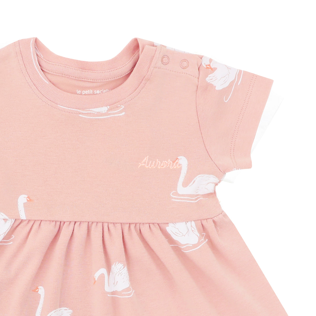 A close-up of a personalised name embroidery on a  peach-coloured, girl's organic dress adorned with a print of swans from Le Petit Society.
