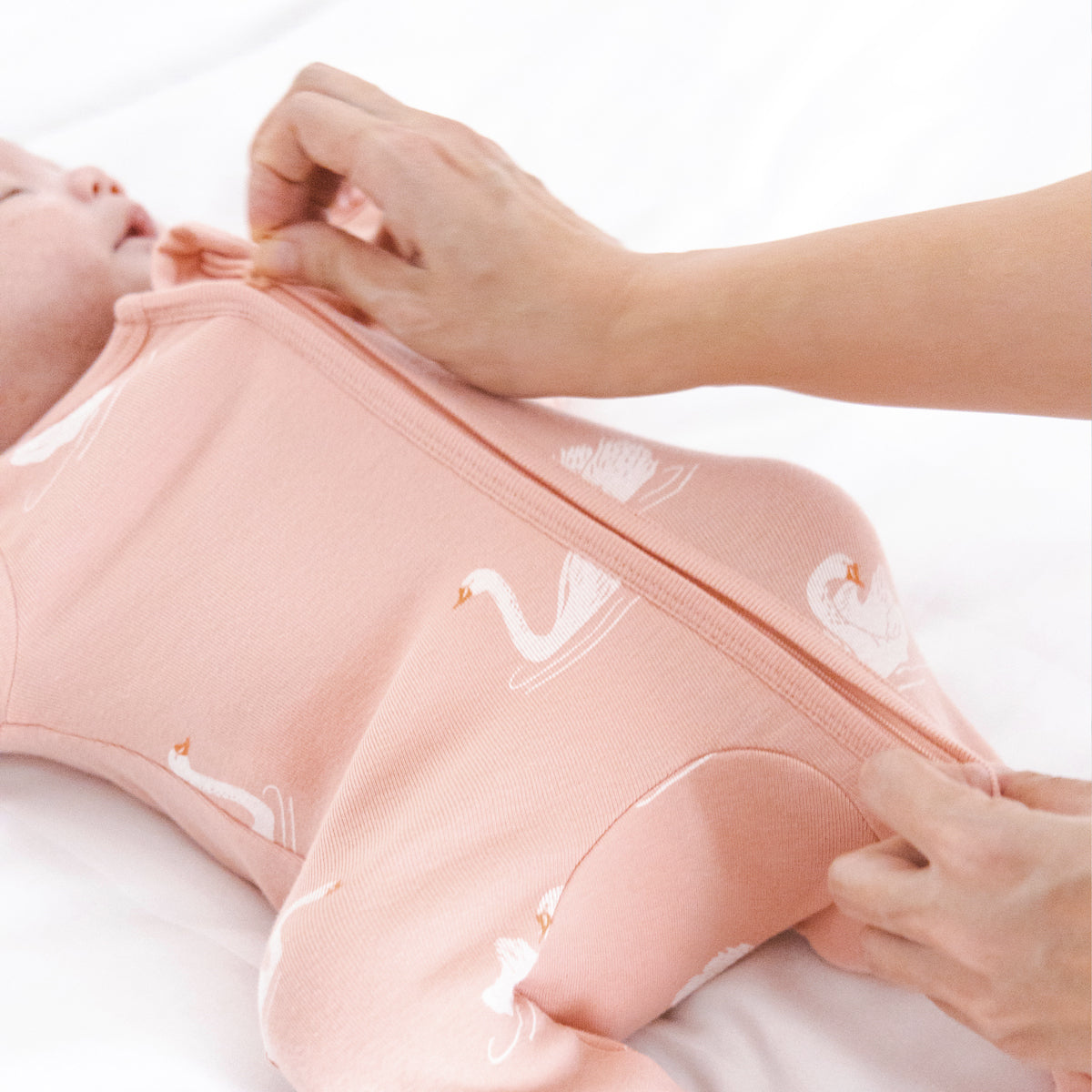Newborn baby being gently changed into a peach-colored organic zip sleepsuit by Le Petit Society, featuring a swan print, foldable mittens, and footies, with the zipper being carefully zipped up.