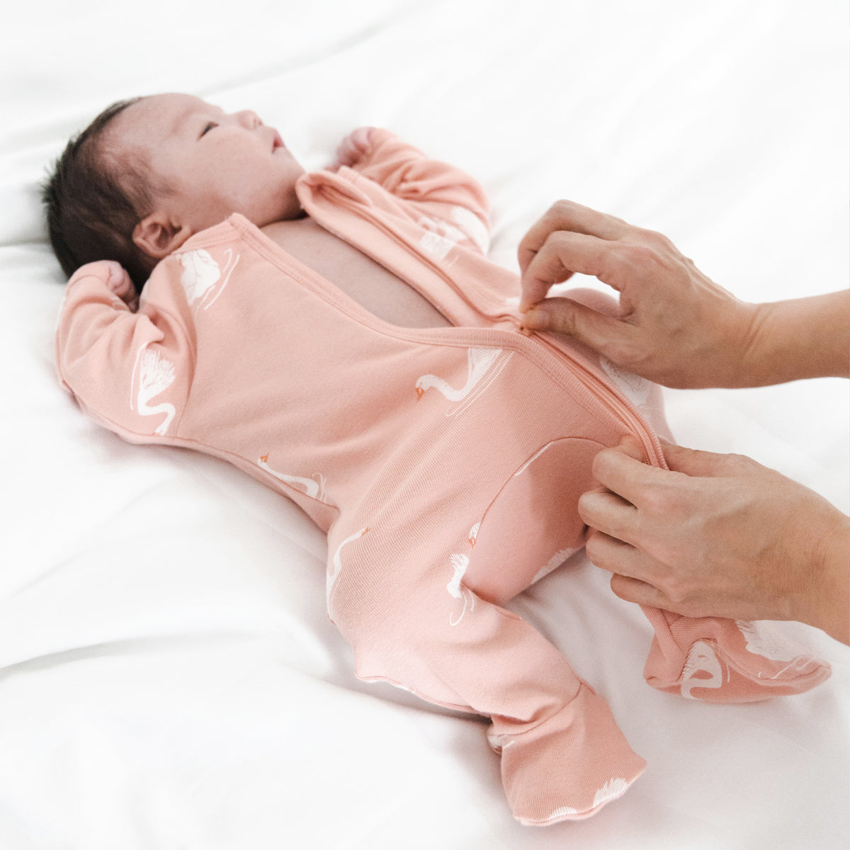 Newborn baby being gently changed into a peach-colored organic zip sleepsuit by Le Petit Society, featuring a swan print, foldable mittens, and footies, with the zipper being carefully zipped up.