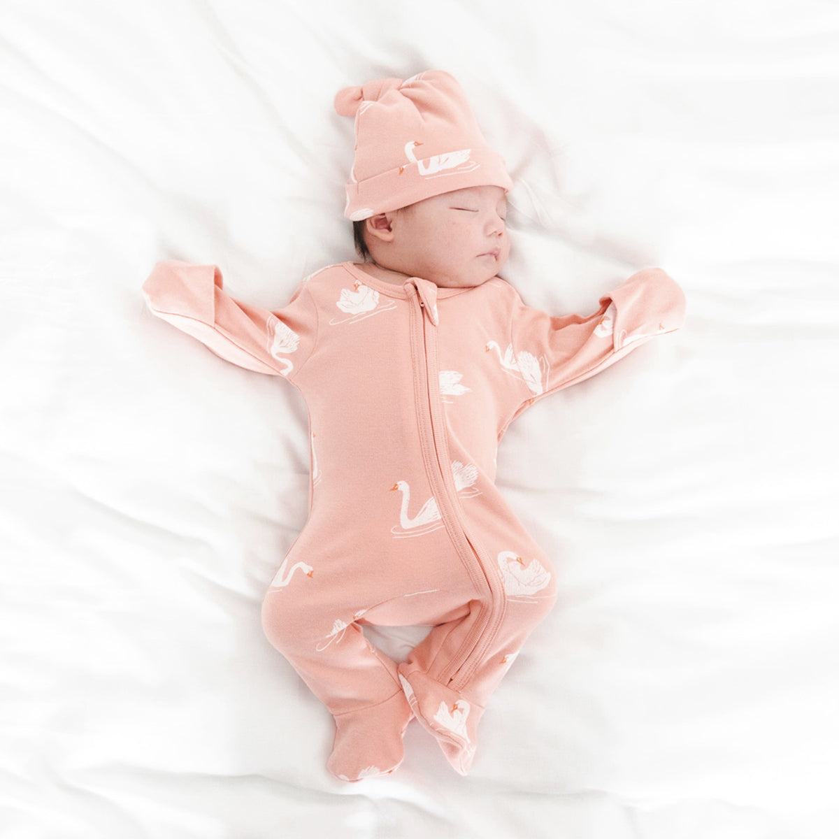 A newborn baby wearing an organic zip sleepsuit in peach with foldable mittens and footies by Le Petit Society, featuring a print of swans.