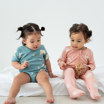 Two little girls wearing pieces from Le Petit Society, one in a kimono romper and the other in an organic zip sleepsuit with foldable mittens and footies, sitting together and sharing snacks.