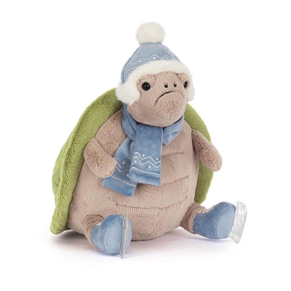 A 45 degree view of a plush turtle toy with a green shell, wearing a blue winter hat with a white pom-pom, a matching blue scarf, and blue skates. The turtle has a soft, beige body and a cute, slightly grumpy expression