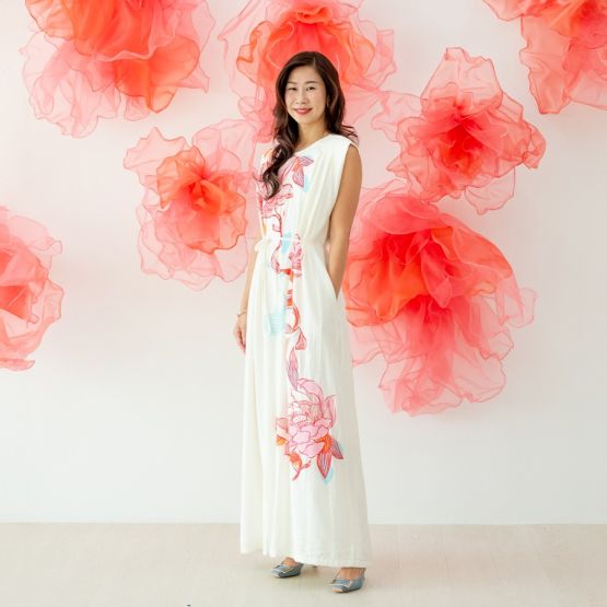 A female model wearing a long cream dress with large pink and blue lotus floral prints from the Le Petit Society x Sheila Sim CNY 2026 collection stands smiling against a white wall decorated with soft coral organza flower accents. She completes the look 