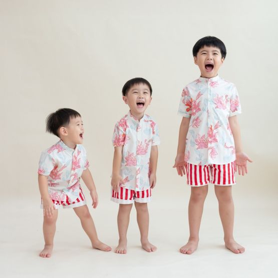 Three boys dressed in matching Lotus Collection outfits stand in a row, bursting into laughter. Their mandarin-collar shirts are covered in coral and pastel lotus illustrations, complemented by bold red-and-white striped shorts. The joyful energy of the m