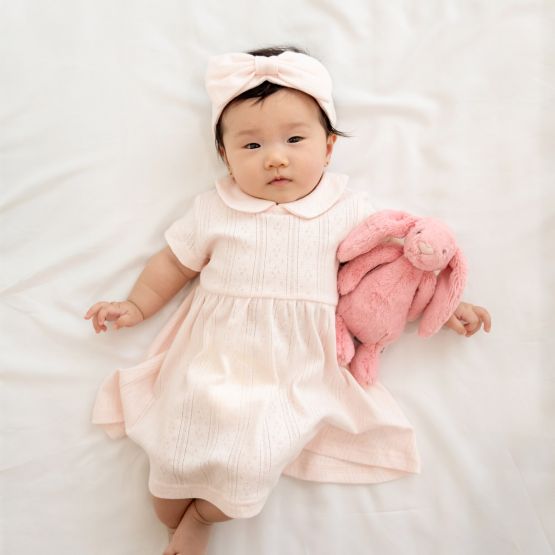 A baby girl wearing a peter pan collar dress in pink pointelle cotton with a matching bow headband.