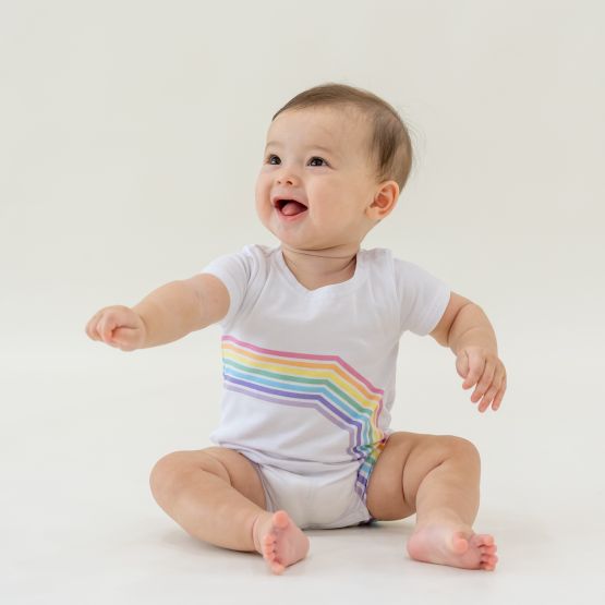 A baby in a white romper with a pastel print of the right arc of a rainbow by Le Petit Society.