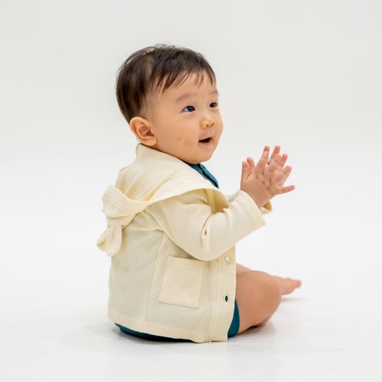 A baby boy wearing a bear cardigan in cream waffle jersey, which comes with buttons and pockets at the front, as well as a hood with little bear ears.