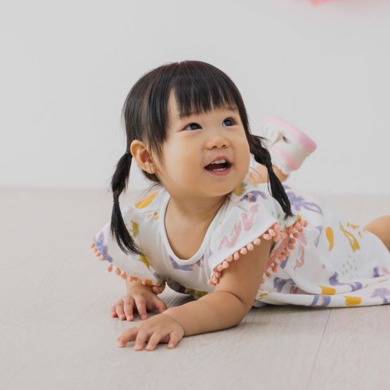 White baby girl’s flutter-sleeve dress from Le Petit Society with pink pompom trim and colorful dragon playground print and sun print in pastel blue, yellow, green, and pink.