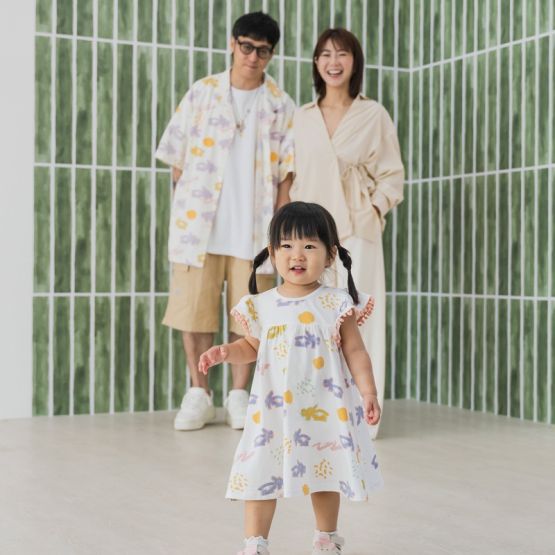White baby girl’s flutter-sleeve dress from Le Petit Society with pink pompom trim and colorful dragon playground print and sun print in pastel blue, yellow, green, and pink.