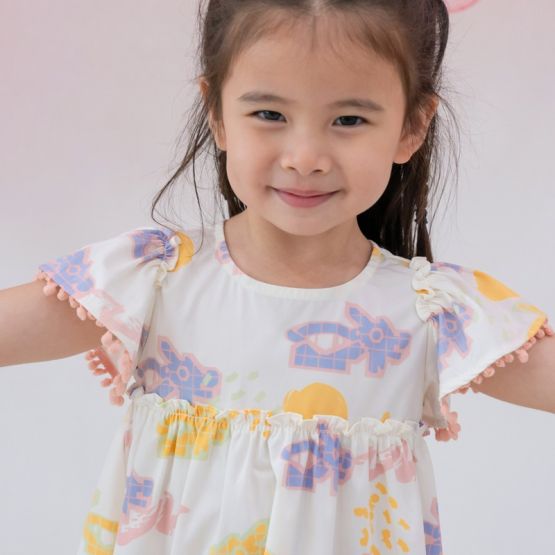 Close-up of white baby girl’s flutter-sleeve dress from Le Petit Society with pink pompom trim and colorful dragon playground print and sun print in pastel blue, yellow, green, and pink.