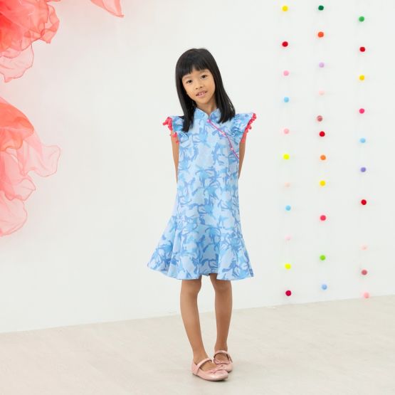 A young girl wearing a light blue ruffle cheongsam dress with pink pom-pom trim from the Le Petit Society x Sheila Sim CNY 2026 collection stands with her hands behind her back. She is smiling softly against a bright backdrop decorated with multicoloured 