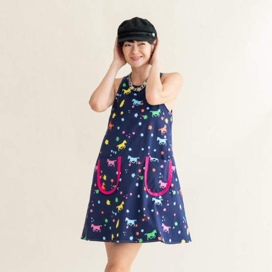 A woman adjusting her black cap while wearing the sleeveless navy dress from the Le Petit Society x Sheila Sim CNY 2026 Rainbow Horse Collection, featuring bright rainbow horse and splatter prints.