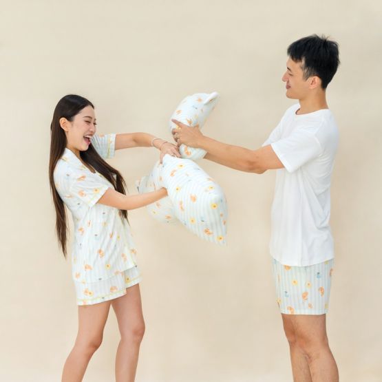 A man and woman having a playful pillow fight, both dressed in matching sleepwear from Le Petit Society featuring a whimsical breakfast food print, with pillows in the same design.