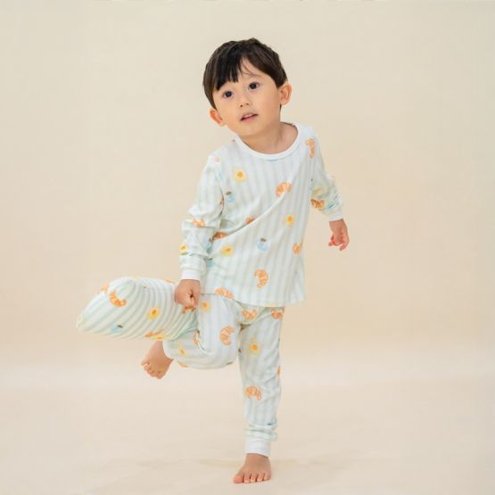 A little boy wearing a pyjama set and holding a matching pillow from Le Petit Society. These pieces feature pastel green stripes and a print of breakfast foo.