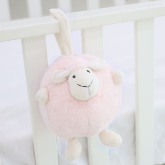 A round and plush sheep toy in soft pink by Le Petit Society hangs off the frame of a baby cot.
