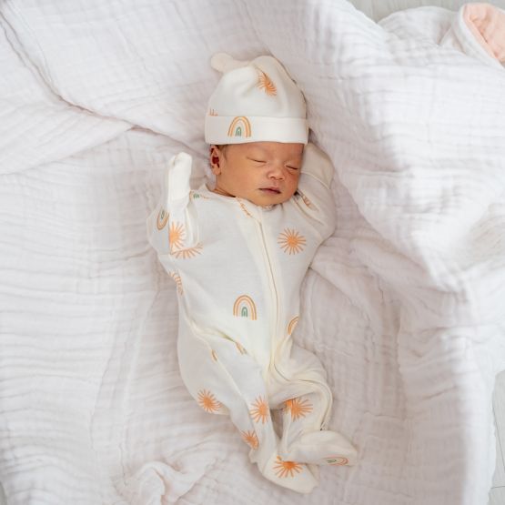A newborn baby wearing an organic zip sleepsuit with foldable mittens and footies by Le Petit Society, featuring a print of sun and rainbows over a cream base. 